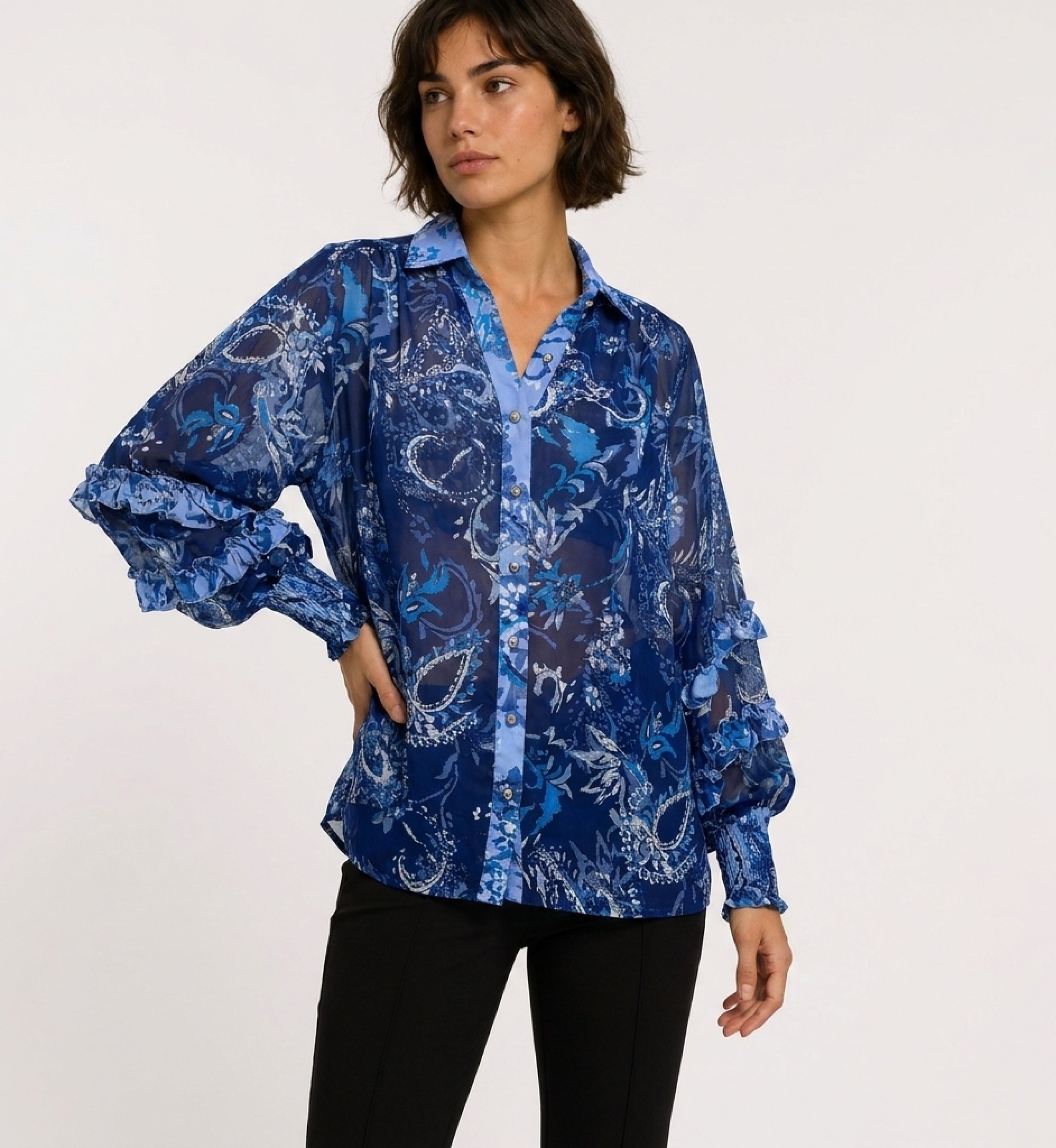 Vista tre quarti di blusa trasparente blu a maniche lunghe con fantasia floreale paisley, polsini a balze e scollo a V.