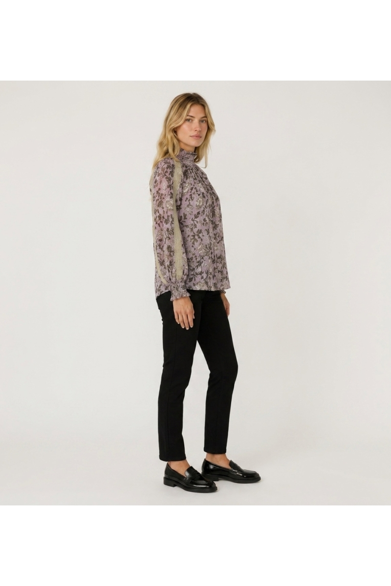 Three-quarter view of a purple long-sleeve high-neck blouse with dark floral pattern, beige lace trim, and smocked cuffs.