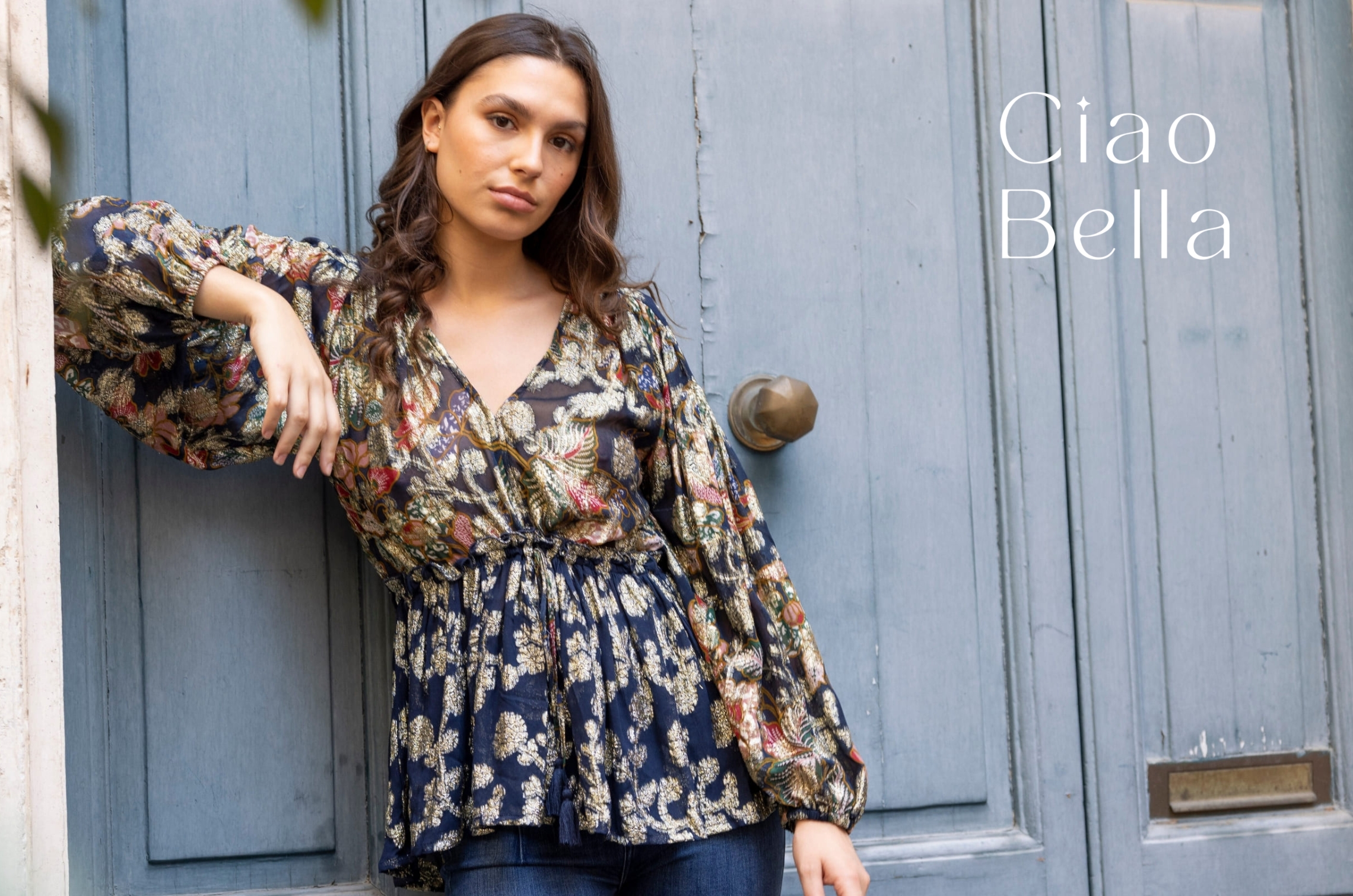 Mannequin portant une blouse à imprimé floral sur fond bleu marine, appuyée contre une porte bleu pastel, avec le texte « Ciao Bella ».